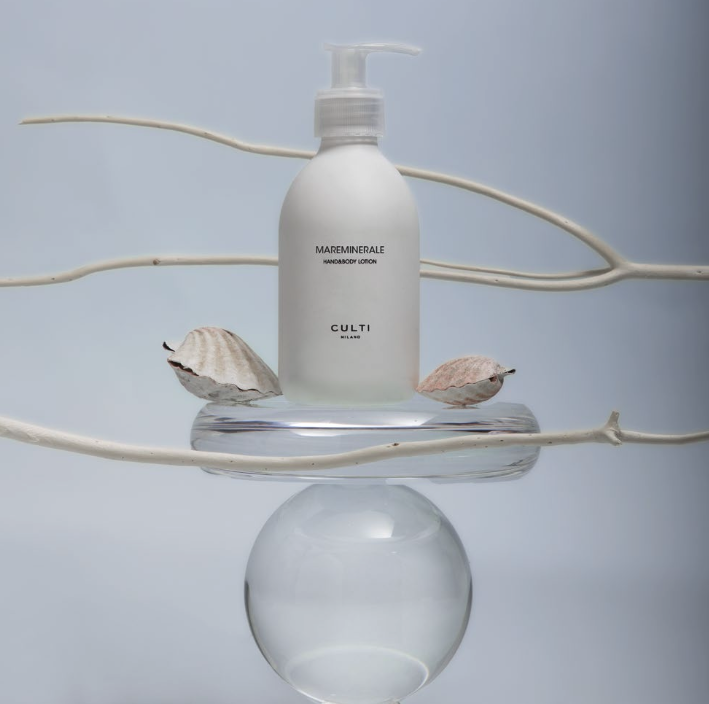 Un flacone bianco di Culti Milano Mareminerale Hand&Body Lotion è bilanciato su vetro trasparente tra rami chiari e conchiglie su sfondo azzurro.