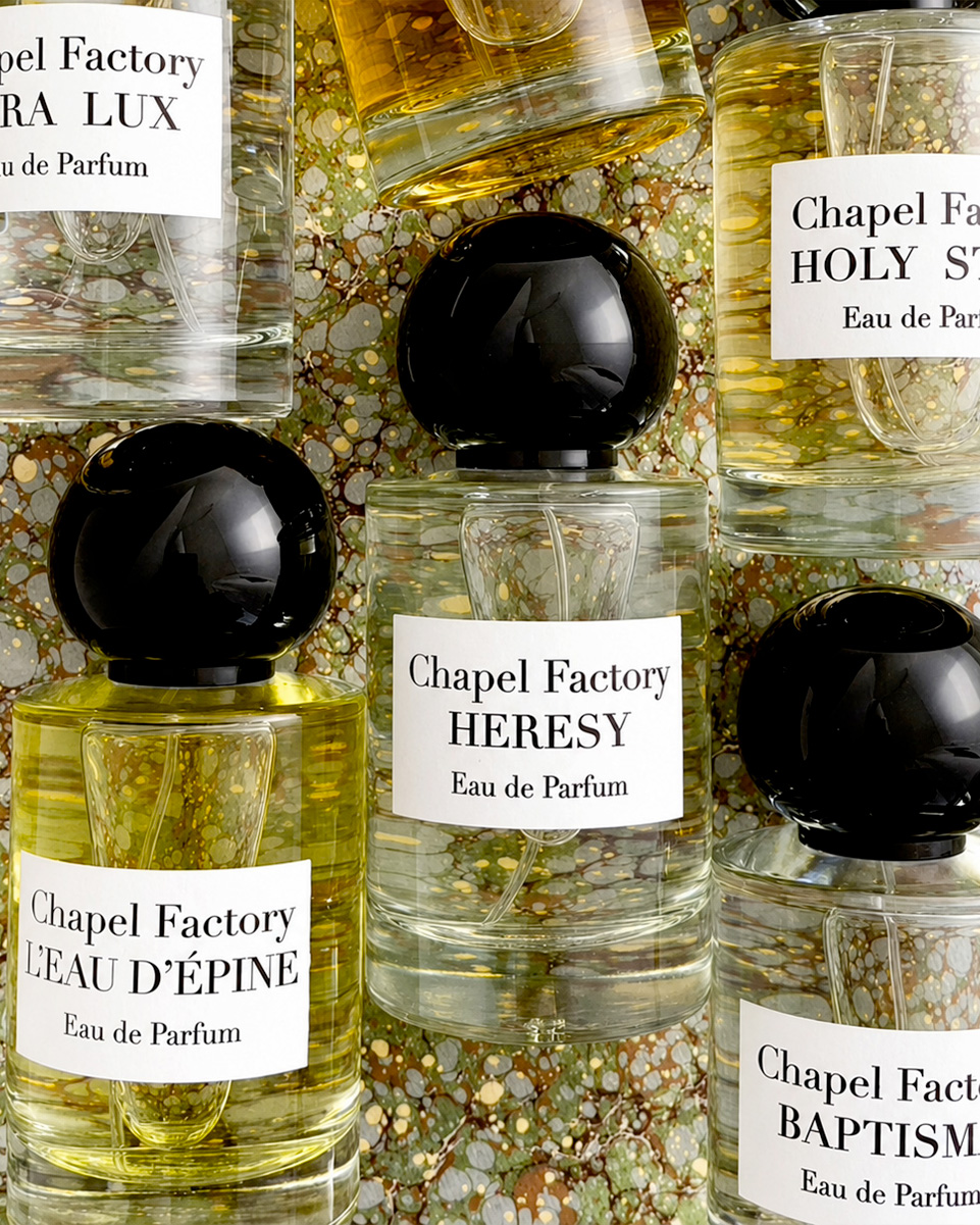 Bottiglie di profumo Chapel Factory con etichette Heresy, Baptism, L’Eau d’Épine e Holy Spirit, disposte su uno sfondo marmorizzato dorato.