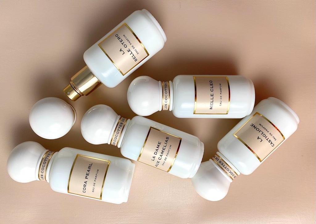 Cinque flaconi di profumo Les Cocottes Paris con etichette dorate sono disposti su fondo beige, con nomi come La Belle Otero e Cora Pearl.