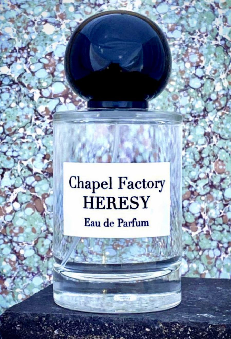 Un flacone di profumo trasparente con tappo sferico nero mostra l'etichetta Chapel Factory HERESY Eau de Parfum davanti a uno sfondo marmorizzato.