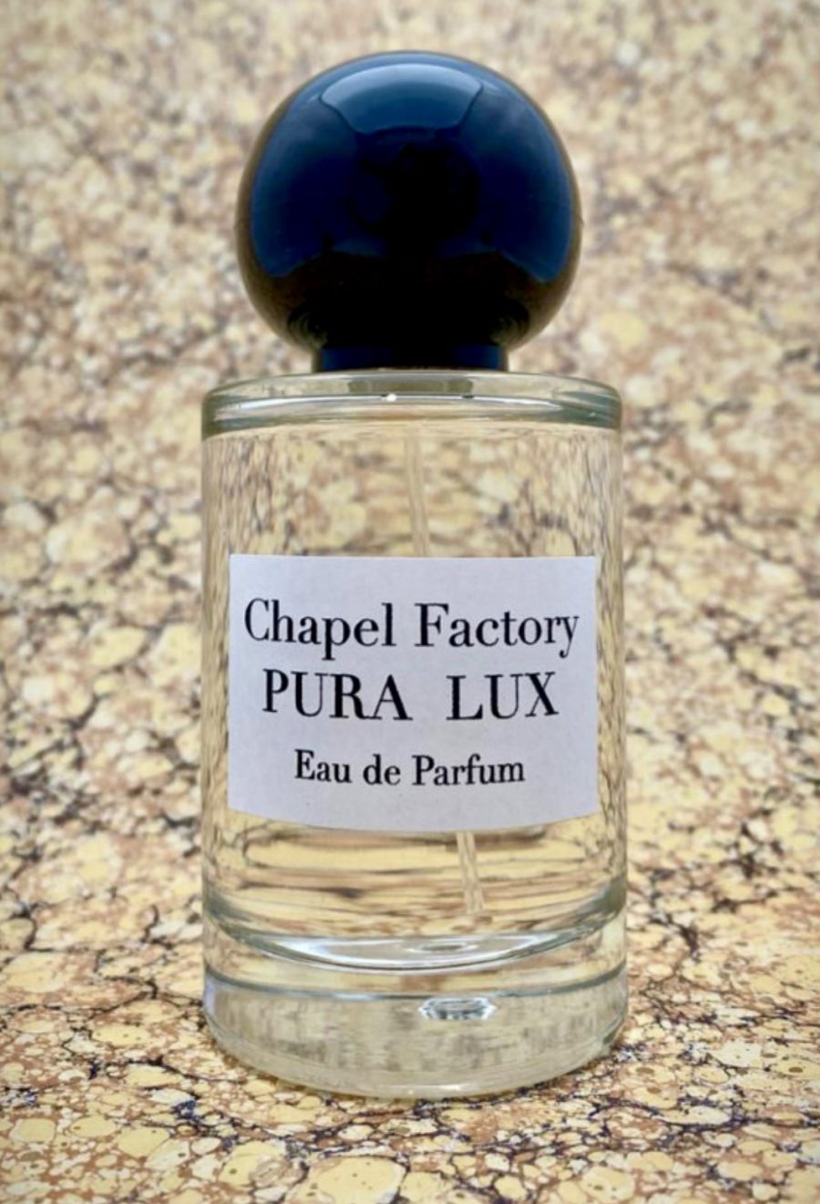 Un flacone di profumo Chapel Factory Pura Lux Eau de Parfum con tappo blu scuro e liquido chiaro, davanti a uno sfondo marmorizzato beige.