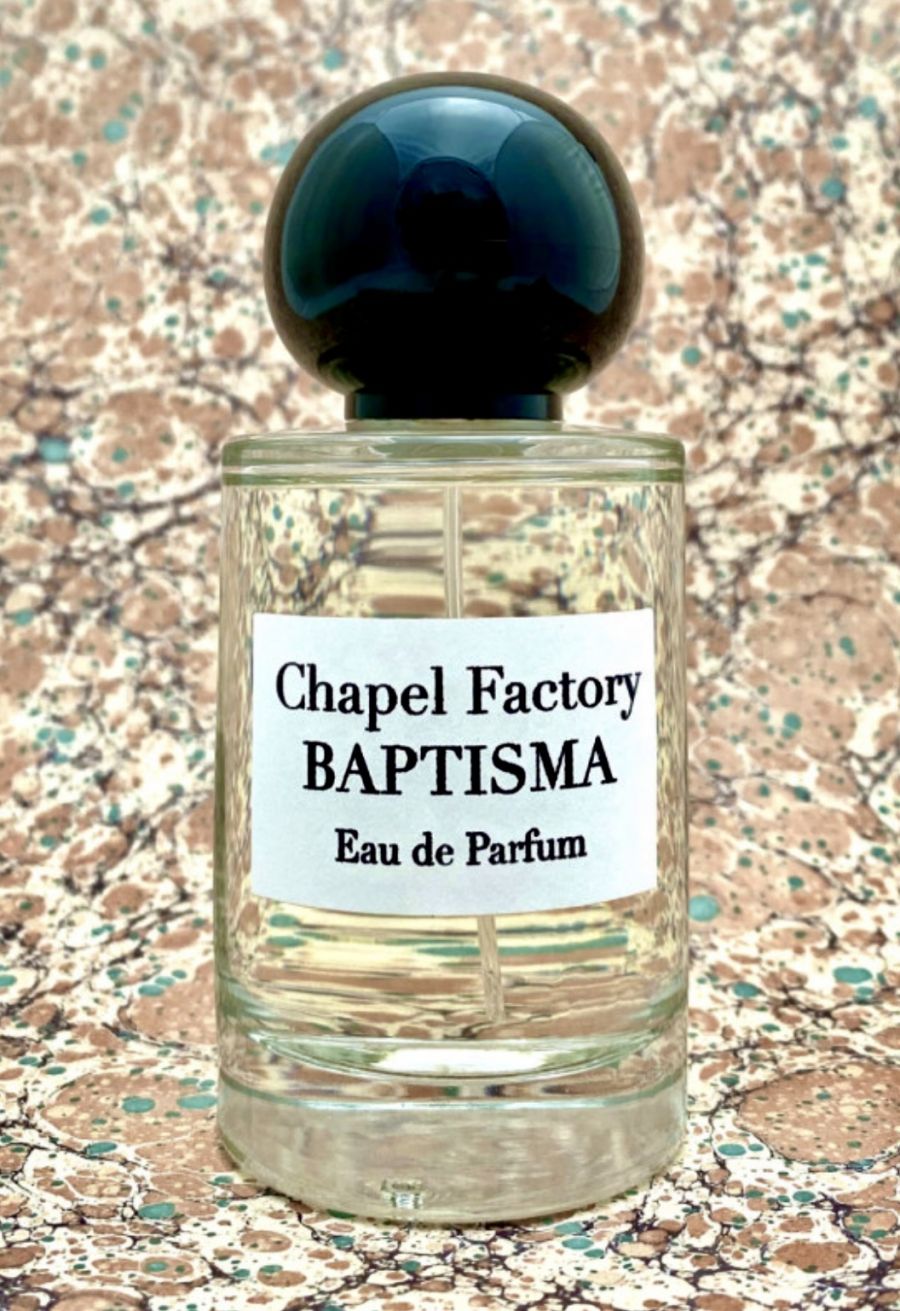 Un flacone di profumo Chapel Factory Baptisma Eau de Parfum con tappo blu scuro e liquido chiaro, davanti a uno sfondo marmorizzato beige.